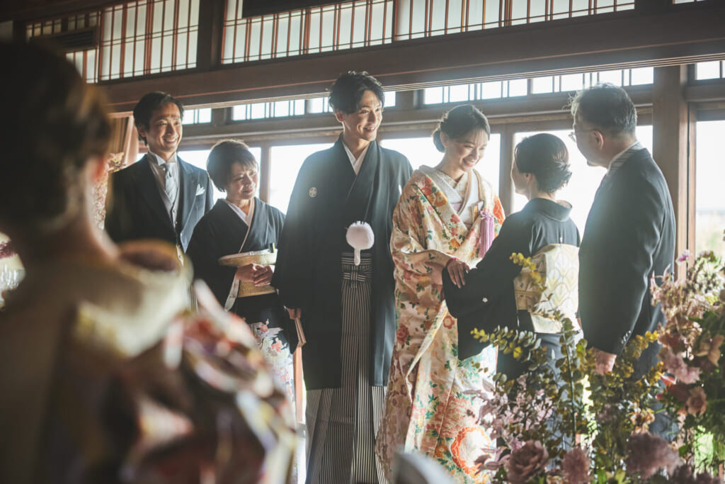 和装の新郎新婦と、両家の家族が室内の窓辺で向き合い、和やかに談笑している集合写真。落ち着いた和の空間が、家族の温かさを引き立てています。
